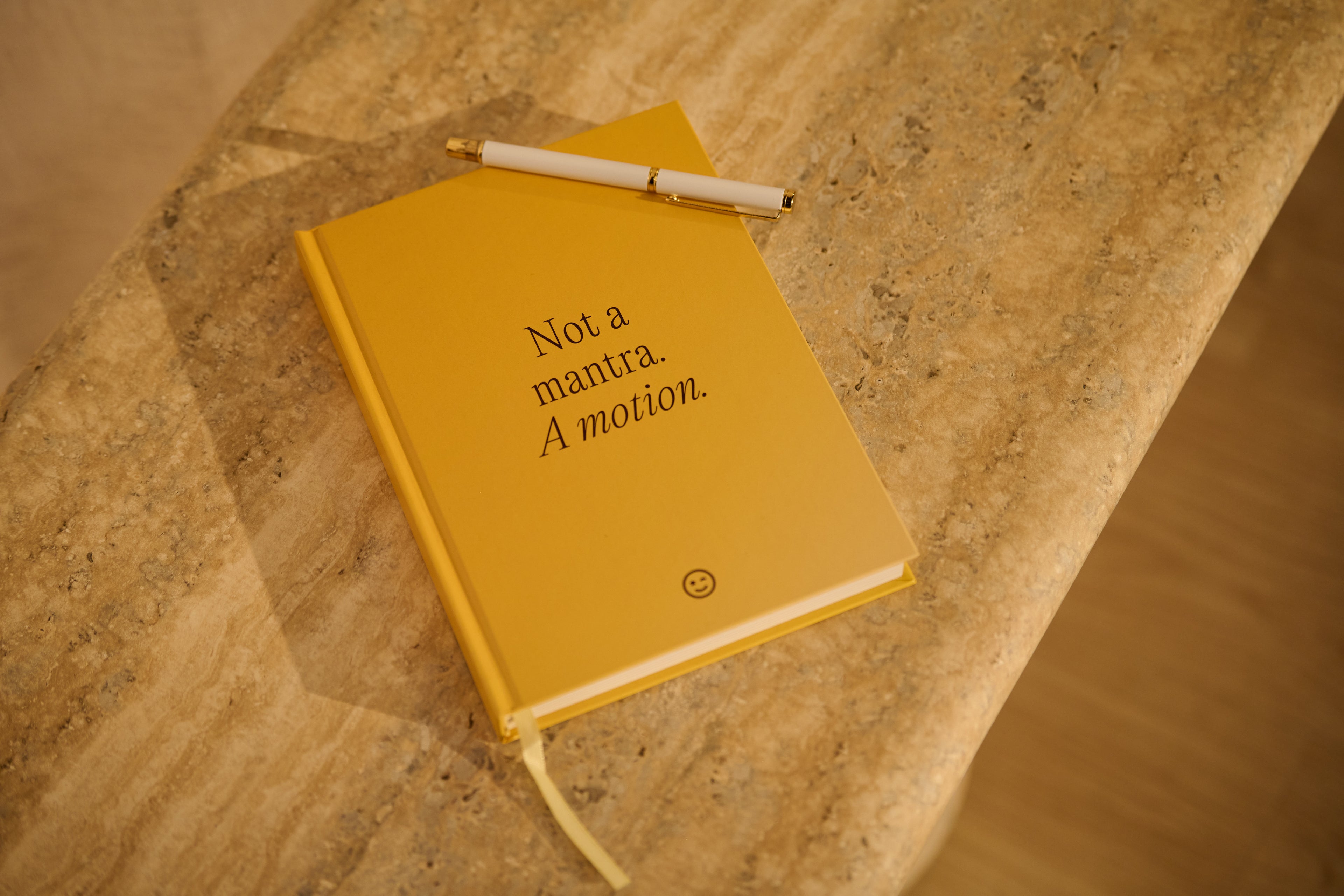 Yellow notebook with a pen on a stone surface, featuring the text 'Not a mantra. A motion.'