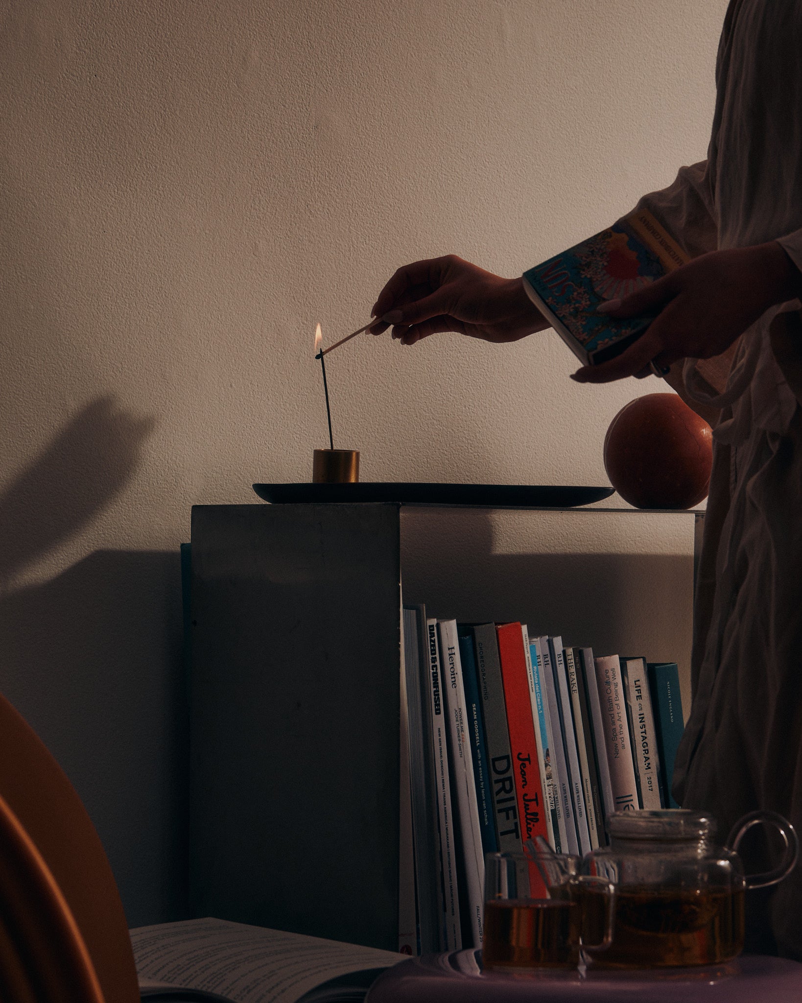 Woman lighting a KIC incense stick on a shelf beside books and loose leaf tea