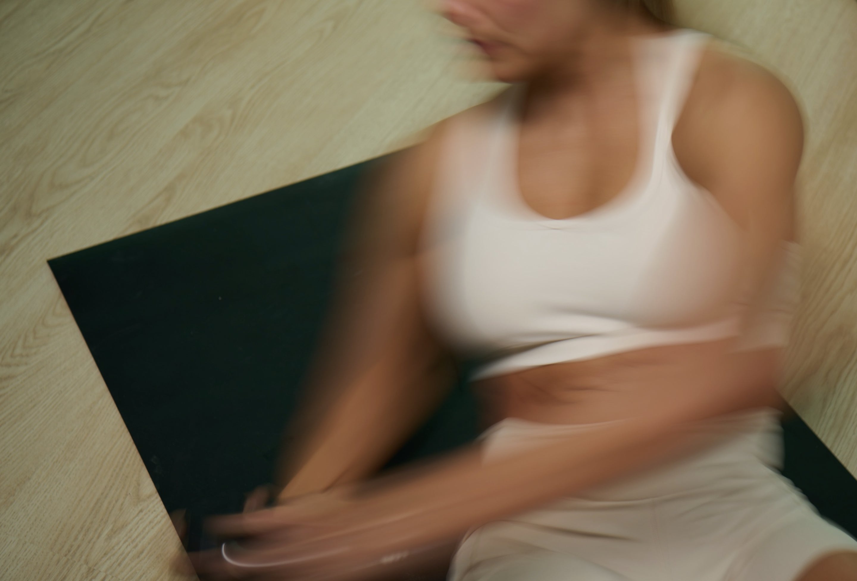 Person in a white tank top doing yoga on a black mat on a wooden floor.