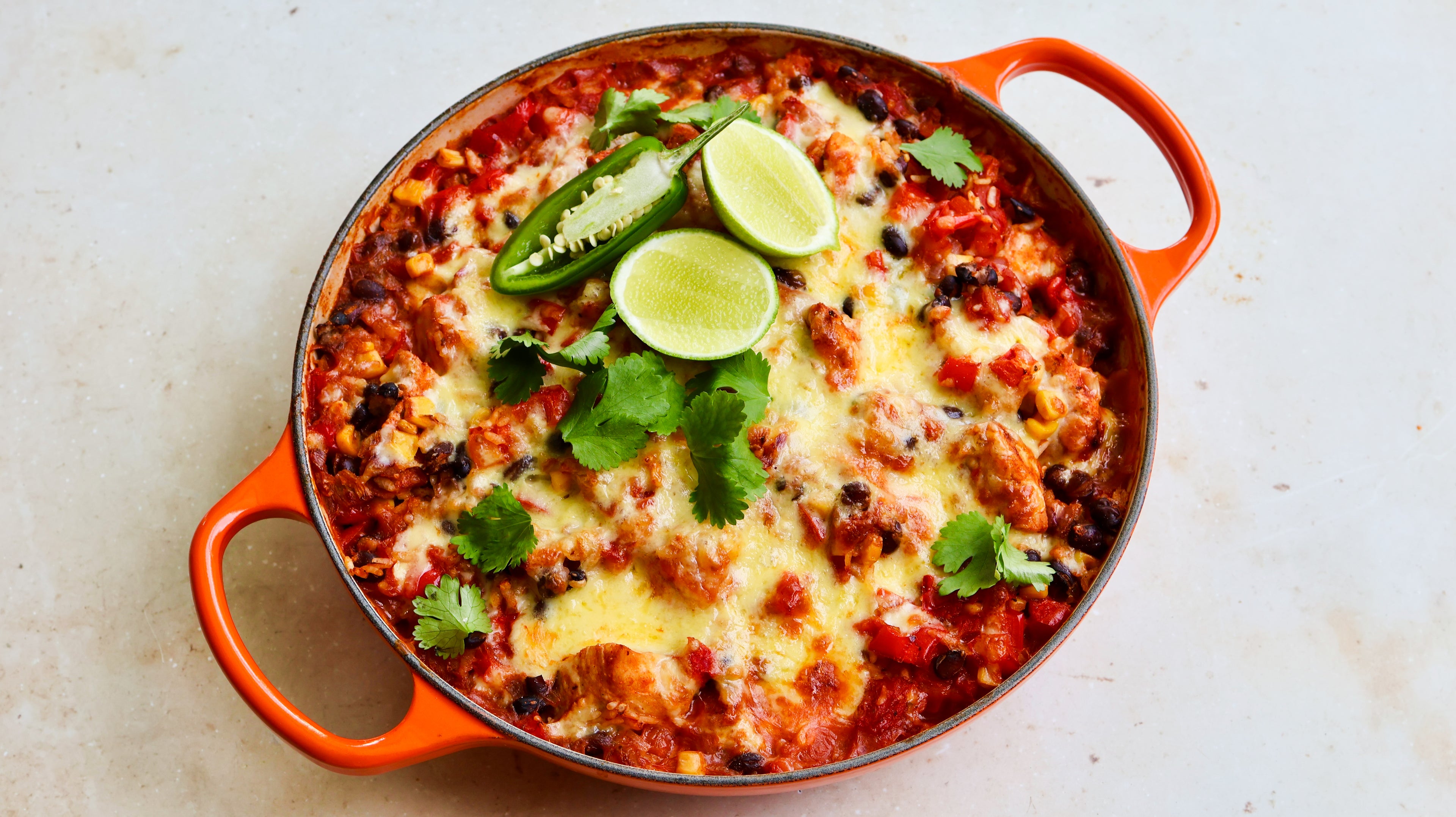 Baked dish with cheese, cilantro, and lime wedges in an orange baking dish on a light background