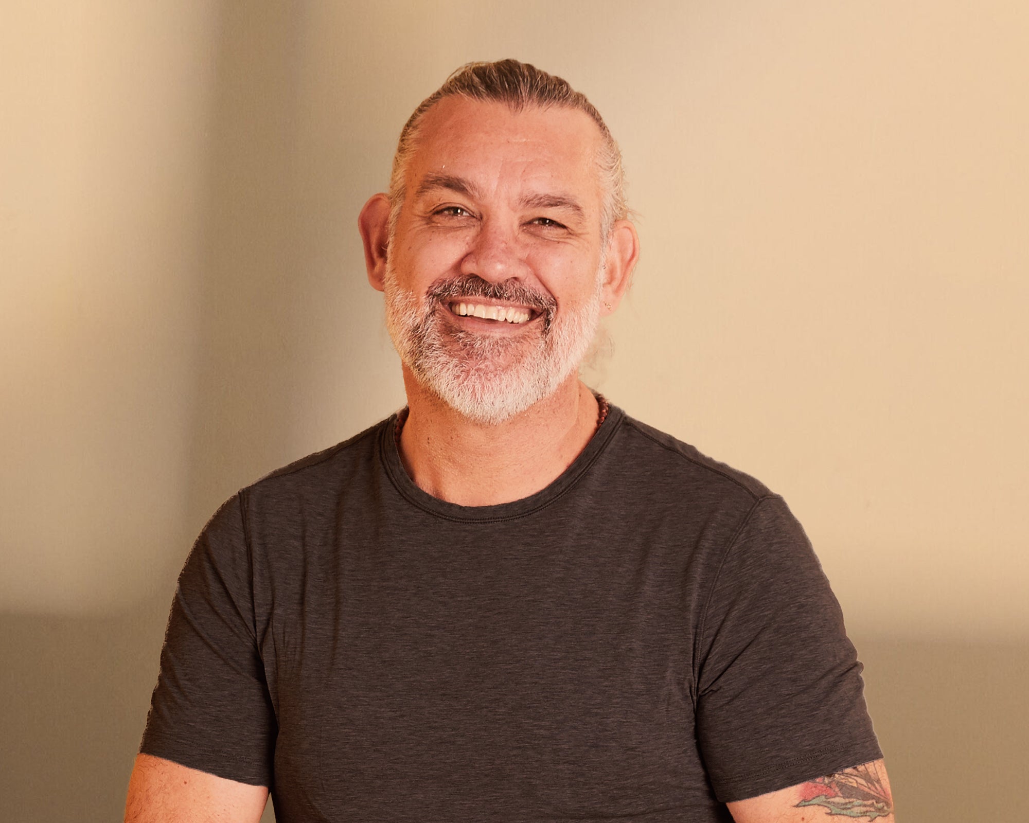 Man with a beard and tattoo on his arm wearing a dark gray t-shirt against a beige background