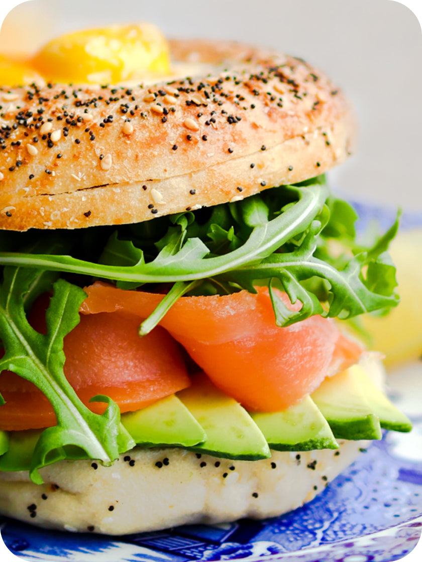 Bagel sandwich with salmon, avocado, and arugula on a blue and white plate.