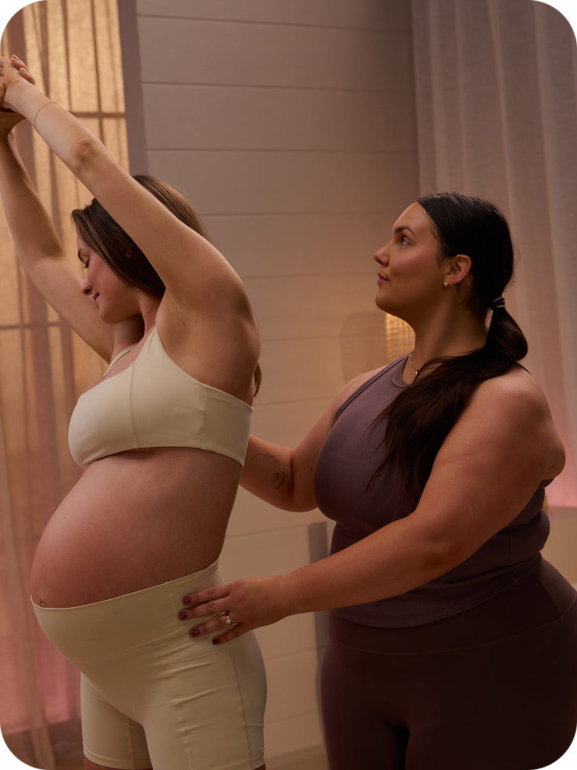 Two women, one pregnant, the other assisting her, in a warm-toned room.